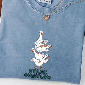 Peut inclure: Un t-shirt bleu jean avec un motif représentant trois oies blanches empilées verticalement. L'oie du haut porte des lunettes et tient un ordinateur portable. Sous les oies, le texte STACK OVERFLOW est écrit en lettres vertes. Étiquette Comfort Colors.