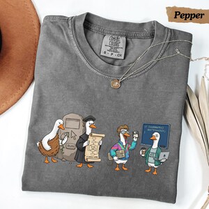 May include: A charcoal gray t-shirt featuring a graphic of four ducks in various tech-related roles. The design includes a duck at a computer, a duck in a graduation cap holding a scroll, a duck with a keyboard, and a duck holding a laptop and coffee.