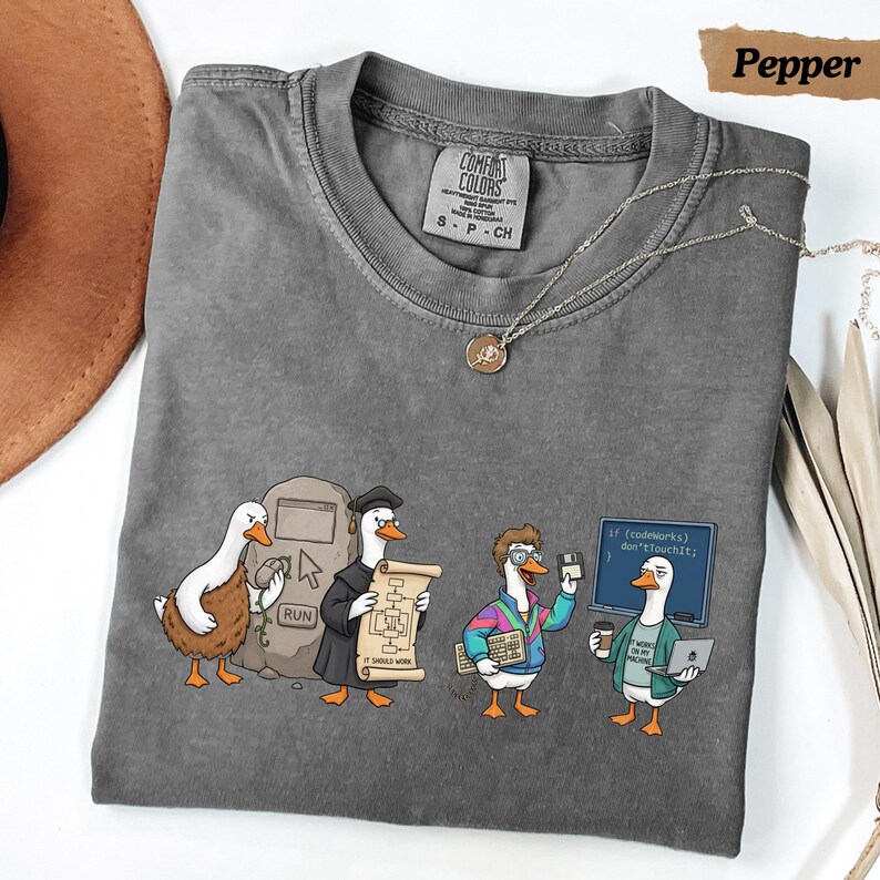 May include: A charcoal gray t-shirt featuring a graphic of four ducks in various tech-related roles. The design includes a duck at a computer, a duck in a graduation cap holding a scroll, a duck with a keyboard, and a duck holding a laptop and coffee.