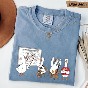 May include: A blue jean coloured t-shirt featuring a graphic of four white geese. One goose points to a whiteboard labelled "WHAT IS ORCHESTRA?" with musical instrument diagrams. Two geese play string instruments, and one wears a red shirt labelled "MAJOR PROBLEM."