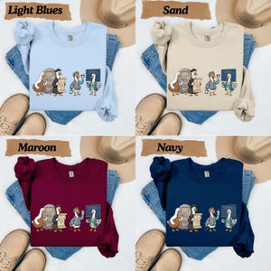 May include: A collection of four crewneck sweatshirts in light blue, sand, maroon, and navy. Each sweatshirt features a graphic print of four ducks in various outfits, including a graduation cap and gown, holding items like a scroll, coffee, and a laptop.