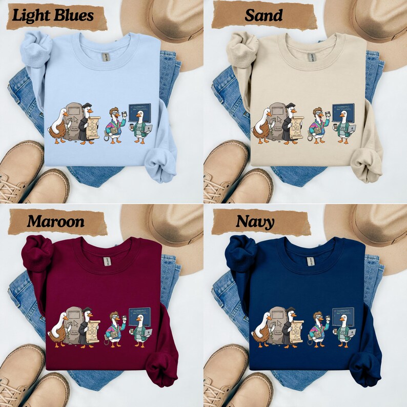 May include: A collection of four crewneck sweatshirts in light blue, sand, maroon, and navy. Each sweatshirt features a graphic print of four ducks in various outfits, including a graduation cap and gown, holding items like a scroll, coffee, and a laptop.