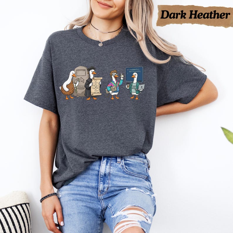 May include: A dark heather gray t-shirt featuring a graphic of four cartoon ducks dressed as different tech professionals. The ducks are depicted with a computer monitor, a scroll, a laptop, and a coffee mug. The text Dark Heather is visible.