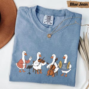 Könnte beinhalten: Ein blaues T-Shirt mit einer Grafik von vier weißen Gänsen, die Musikinstrumente spielen. Eine Gans dirigiert, eine spielt Geige, eine spielt Cello und eine hält Becken, während sie ein Shirt mit der Aufschrift Not My Tempo trägt.