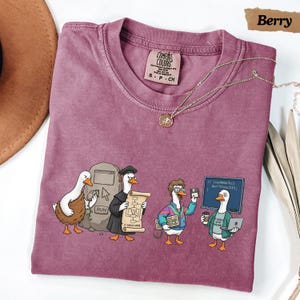 May include: A berry-colored crew neck t-shirt featuring a graphic of four ducks in various tech-related roles. The print includes a duck with a computer mouse, a duck in a graduation cap holding a scroll, a duck with a keyboard, and a duck with a laptop and coffee cup in front of a chalkboard that reads: if (codeWorks) don'tTouchIt; }.