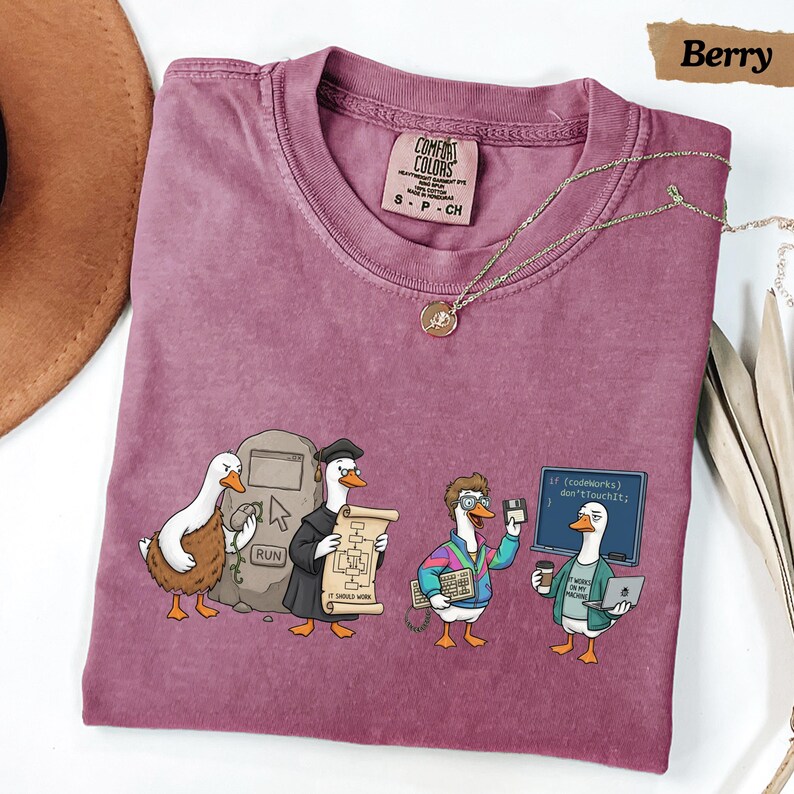 May include: A berry-colored crew neck t-shirt featuring a graphic of four ducks in various tech-related roles. The print includes a duck with a computer mouse, a duck in a graduation cap holding a scroll, a duck with a keyboard, and a duck with a laptop and coffee cup in front of a chalkboard that reads: if (codeWorks) don'tTouchIt; }.