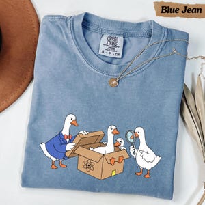 Peut inclure: Un t-shirt bleu jean orné d'un motif représentant trois oies blanches interagissant avec une boîte en carton. Une oie porte un blazer bleu et un nœud papillon rouge, une autre est assise dans la boîte et la troisième tient une loupe.