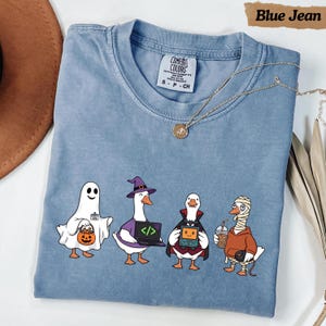 May include: A blue jean colored t-shirt featuring a graphic of four geese dressed in Halloween costumes: a ghost holding a pumpkin bucket, a witch with a laptop, a vampire with a handheld game, and a mummy holding a drink. The shirt has a crew neck.