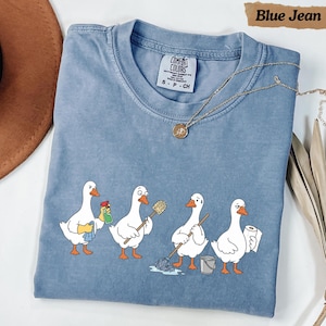 Puede incluir: Una camiseta azul con un gráfico de cuatro gansos blancos realizando tareas de limpieza. Un ganso sostiene una botella de spray, otro un cepillo de baño, otro una fregona y otro un rollo de papel de cocina. La camiseta tiene cuello redondo.