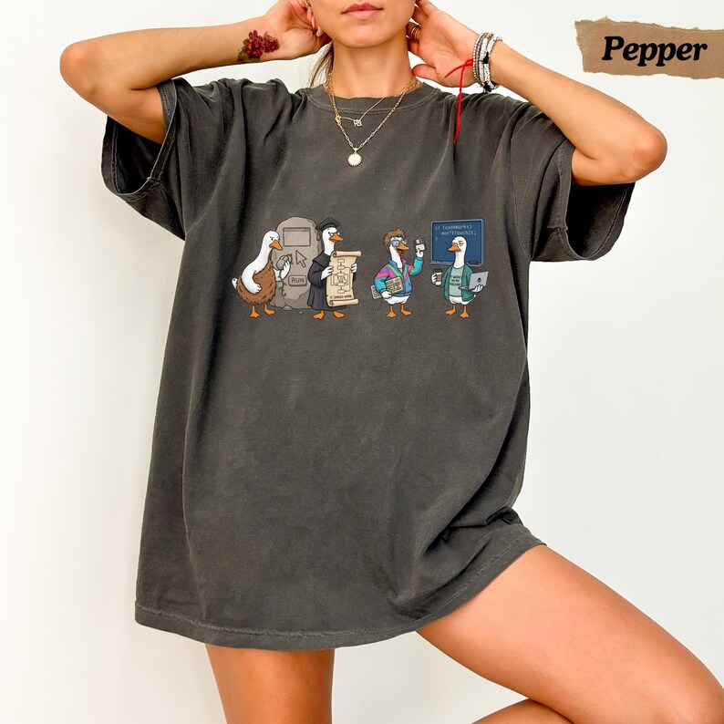 May include: A charcoal gray short-sleeve t-shirt featuring a graphic print of four cartoon ducks in various outfits. The design includes a duck with a computer, a duck holding a scroll, a duck in a colorful jacket, and a duck with a laptop.