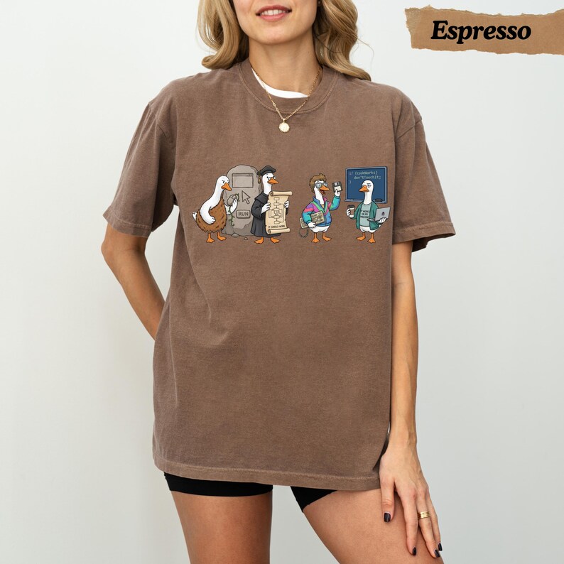 May include: A brown short-sleeve t-shirt featuring a graphic of four cartoon geese in various outfits. The geese are interacting with a computer screen, a scroll, a tablet, and a laptop. The text "Espresso" appears in the top right corner.