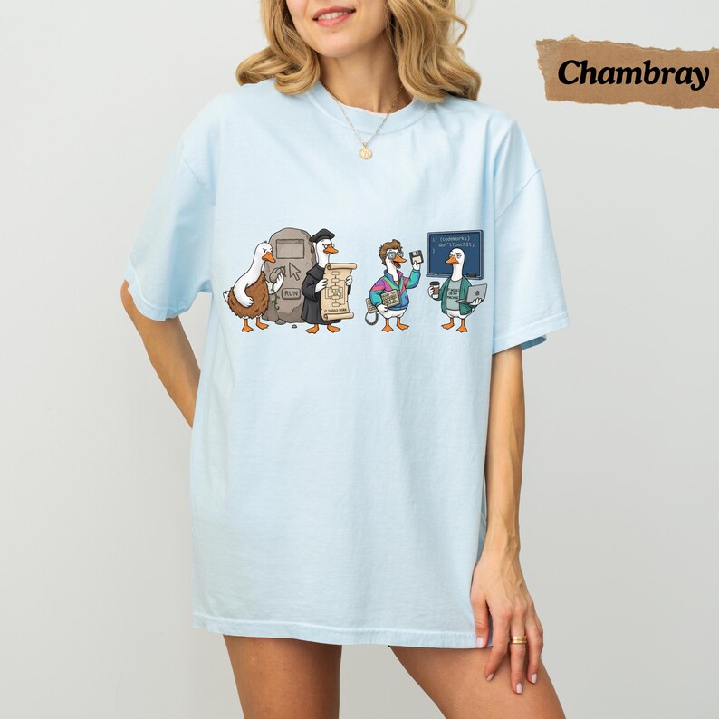 May include: A light blue t-shirt featuring a graphic of four cartoon ducks in various outfits. The ducks are depicted with a stone tablet, a scroll, a floppy disk, and a laptop. The design includes a small chalkboard with text in the background.