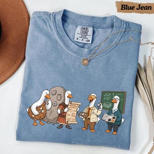 Peut inclure: Un t-shirt bleu avec un motif représentant quatre canards blancs illustrant différentes époques de l'histoire et de la science. Les canards sont représentés en homme des cavernes, en Romain, en scientifique en blouse et en chercheur moderne.