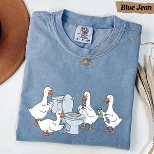 May include: A blue jean colored t-shirt featuring a graphic of four white geese cleaning a toilet. One goose holds a toilet brush, another a spray bottle, and two others hold cleaning cloths. The shirt has a Comfort Colors brand label.