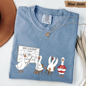 May include: A blue jean coloured t-shirt featuring a graphic of four white geese in a classroom setting. One goose stands at a whiteboard with the text "WHAT IS ALGEBRA?" and various math equations, while the others sit or stand, one wearing a red shirt with "ASK Y?" printed on it.