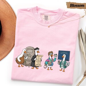 May include: A light pink short-sleeve t-shirt featuring a graphic of four ducks dressed as computer programmers. The illustration includes a duck with a stone tablet, a duck in a graduation gown holding a scroll, a duck with glasses, and a duck with a laptop and coffee cup. The text on the screen reads: if (codeworks) don'tTouchIt;.