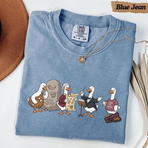 Könnte beinhalten: Ein blaues T-Shirt mit Rundhalsausschnitt und einer Grafik von vier weißen Enten als Musiker. Eine Ente hält einen Knochen, eine andere eine Schriftrolle mit der Aufschrift Orchestra, eine dirigiert und eine trägt ein Shirt mit Stay In Tune.