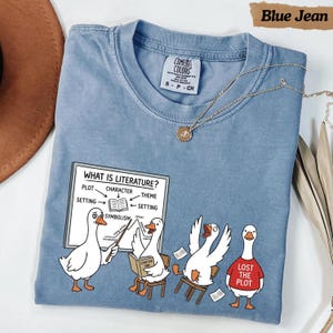 May include: A blue jean colored t-shirt featuring a graphic of four white geese in a classroom setting. One goose points to a whiteboard labeled "WHAT IS LITERATURE?" with a diagram. Another goose wears a red shirt with the text "LOST THE PLOT."