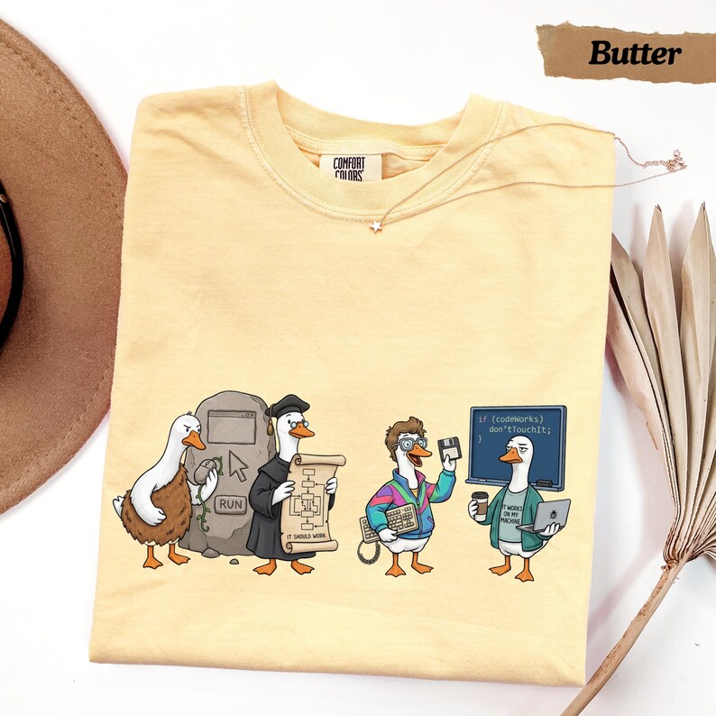 May include: A butter-colored t-shirt featuring a graphic of four ducks in various tech-themed roles. One duck stands by a stone tablet with a run button, another wears a graduation cap holding a flowchart, a third holds a keyboard and floppy disk, and the last holds a laptop and coffee in front of a chalkboard with the text: if (codeworks) don'tTouchIt;.