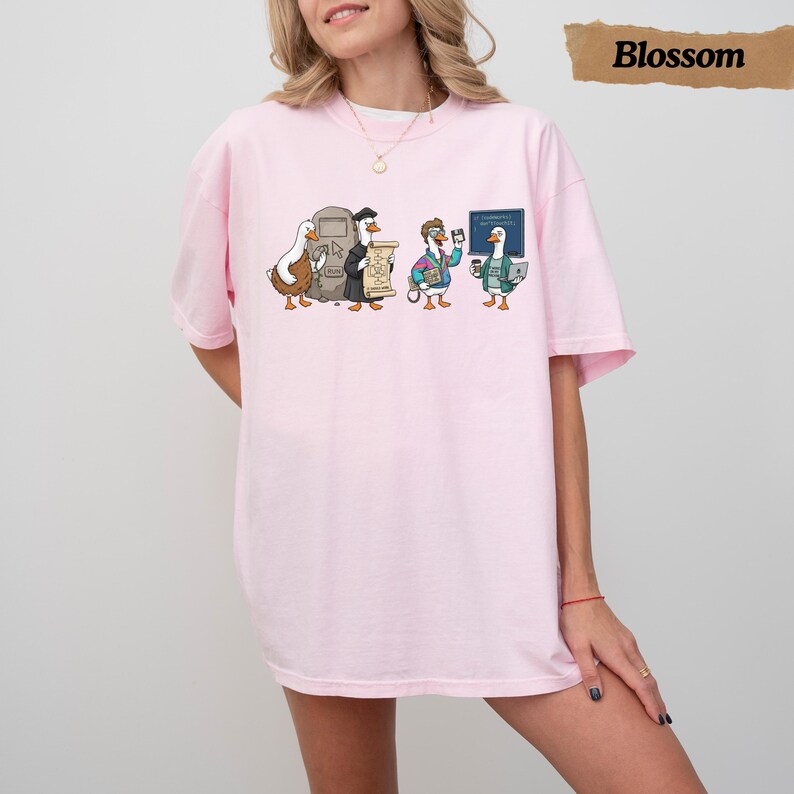 May include: A light pink short-sleeve t-shirt featuring a graphic print of four cartoon ducks dressed in various outfits. The ducks are interacting with a stone tablet, a scroll, a floppy disk, and a laptop. The design includes a blue chalkboard.