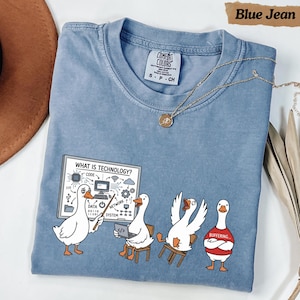 Peut inclure: Un t-shirt bleu jean avec un motif représentant quatre oies blanches. Une oie pointe un tableau blanc intitulé WHAT IS TECHNOLOGY? avec diverses icônes. Une autre utilise un ordinateur portable, une est assise ailes levées, et une porte un t-shirt rouge marqué BUFFERING.