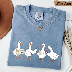Pode incluir: Uma t-shirt azul cor de ganga com um gráfico de quatro gansos brancos. Um ganso segura uma placa que diz "HAND + SCHUH" com ilustrações de uma luva e um sapato. Os outros gansos interagem com uma luva e um sapato de cor castanha.