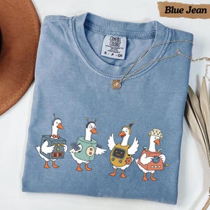 May include: A blue jean coloured t-shirt featuring a graphic of four white geese dressed in various robot costumes with antennas and control panels. The shirt has a crew neckline and a Comfort Colors brand label on the inside of the collar.