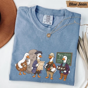 Puede incluir: Una camiseta de color azul vaquero con un gráfico de cuatro patos antropomórficos vestidos de científicos y exploradores. Los patos aparecen con una losa de piedra, un pergamino de zoología, binoculares y un marco con cráneos de animales.