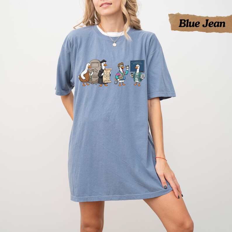 May include: A blue jean colored t-shirt featuring a graphic print of four cartoon geese. The geese are depicted in various outfits, including one wearing a graduation cap and gown, and another holding a floppy disk. The design includes a small chalkboard.