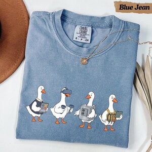 May include: A blue jean coloured t-shirt featuring a graphic of four white ducks. One duck wears a vest and holds a tablet, one wears a VR headset with controllers, one wears a boxy metallic suit, and one wears a utility harness with a circuit board.