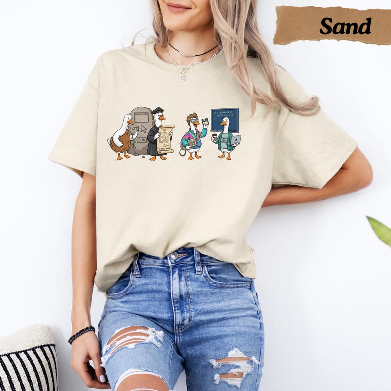 May include: A sand-colored short-sleeve t-shirt featuring a graphic print of four ducks evolving through history. The design shows a prehistoric duck, a medieval duck with a scroll, a 1980s-style duck with a floppy disk, and a modern duck with a laptop.