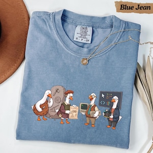 Puede incluir: Una camiseta azul de cuello redondo con un estampado gráfico de cuatro gansos que evolucionan a través de la historia, desde un ganso prehistórico con una herramienta de piedra hasta uno moderno con una placa de circuito. Etiqueta Comfort Colors.