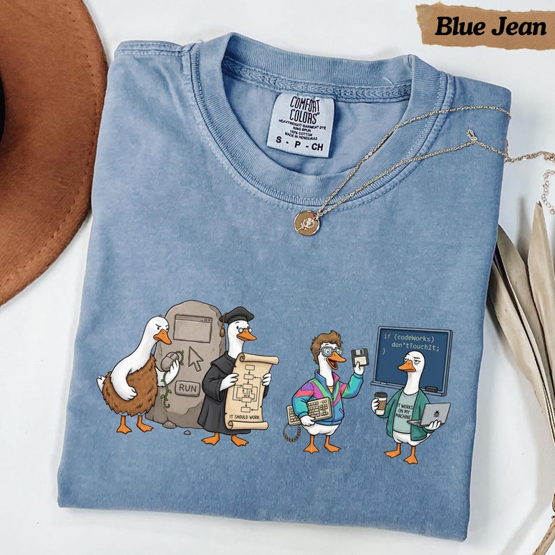 May include: A blue short-sleeve t-shirt featuring a graphic of four ducks dressed as computer programmers. The design includes a stone tablet with a run button, a duck in a robe holding a scroll, a duck with a keyboard, and a duck with a laptop.