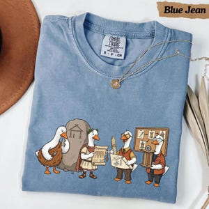 Könnte beinhalten: Ein blaues Kurzarm-T-Shirt mit einer Grafik von vier Enten, die verschiedene Epochen der Ingenieurskunst darstellen. Die Enten sind mit Werkzeugen, einer Steintafel und Bauplänen abgebildet. Das T-Shirt hat einen Rundhalsausschnitt und ein Comfort Colors Etikett.