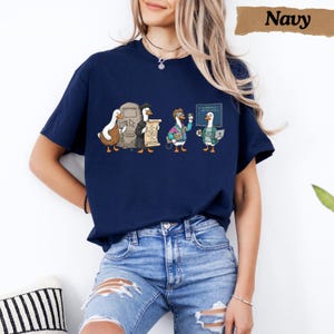 May include: A navy blue t-shirt featuring a graphic print of four cartoon ducks in various roles, including a student with a scroll, a person with glasses, and a duck holding a laptop in front of a chalkboard with code text written on it.
