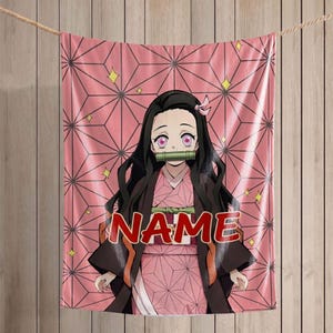May include: A rectangular fleece blanket featuring a pink and black geometric pattern background with a central anime character illustration. The character has long black hair, pink eyes, and a green bamboo muzzle. The word NAME is printed in red.