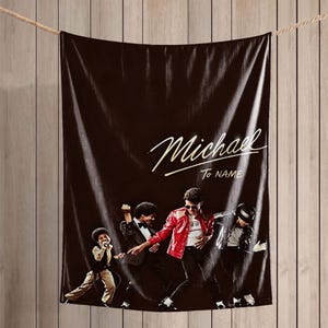 May include: A black rectangular throw blanket featuring a graphic print of four figures in various dance poses. The text Michael is printed in a large script font above the words To NAME in a smaller sans-serif font on the upper right side.
