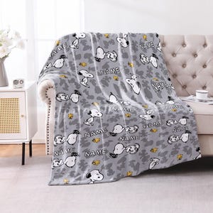 May include: A gray fleece throw blanket featuring a repeating pattern of Snoopy and Woodstock characters in black and white outlines, interspersed with the word NAME printed in black capital letters against a subtle gray camouflage background.
