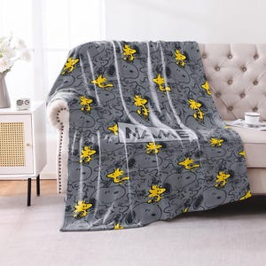 May include: A gray fleece blanket featuring a repeating pattern of Snoopy and Woodstock characters in yellow and black outlines. A rectangular white box is centered on the blanket with the word NAME printed in bold black capital letters.