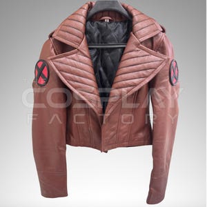 May include: A brown leather jacket featuring wide, quilted lapels and long sleeves. Each shoulder displays a circular patch with a black X on a red background. The interior is lined with black quilted fabric, and the jacket is displayed on a hanger.