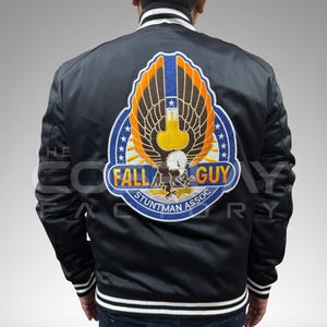 May include: A black bomber jacket with white striped ribbed collar, cuffs, and hem. The back features a large embroidered patch of an eagle with orange and brown wings, blue accents, and the text FALL GUY STUNTMAN ASSOC in bold yellow letters.