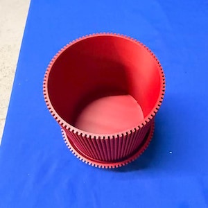 May include: A cylindrical red container featuring a deeply ribbed exterior texture and a prominent gear-like notched rim along the top and bottom edges. The interior is smooth and hollow, showcasing a consistent matte red finish throughout the object.