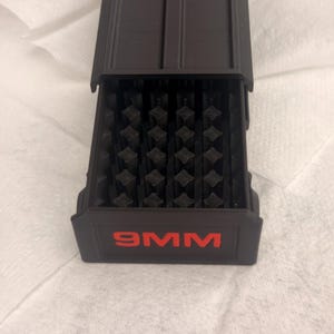 May include: A black rectangular ammunition storage box with a sliding drawer featuring a grid of individual slots for cartridges. The front of the drawer is clearly marked with the text 9MM in bold red lettering against the matte black surface.