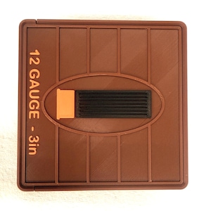 May include: A square brown plastic gauge tool featuring vertical embossed lines and a central oval cutout. The text 12 GAUGE - 3in is printed in orange on the left side. A small orange and black rectangular component is centered in the oval.