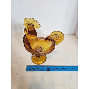 May include: An amber-colored glass rooster figurine with textured feather details, standing on a circular base. A blue ruler is positioned in front, showing the figurine measures approximately 7 inches in length from beak to tail.