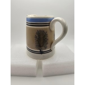 May include: A ceramic pitcher featuring a light blue band at the top, followed by a tan middle section decorated with dark brown tree-like dendrite patterns. The base is white, and the pitcher includes a curved handle and a small pouring spout.