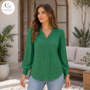 Può includere: Una camicetta verde smeraldo a maniche lunghe con scollo a V e delicati dettagli in pizzo lungo il colletto e sul centro davanti. Il tessuto presenta una sottile trama plissettata verticale e le maniche sono rifinite con polsini aderenti.