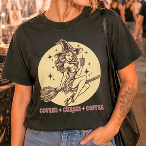 May include: A black t-shirt featuring a graphic of a witch with long wavy hair wearing a pointed hat, riding a broomstick while holding a coffee cup against a large pale yellow moon. The text COVENS + CURSES + COFFEE is printed in pink below.