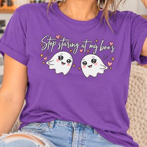 Pode incluir: Uma t-shirt roxa de manga curta com uma estampa gráfica de dois fantasmas brancos sorridentes com bochechas rosadas. O texto acima dos fantasmas diz, Stop staring at my boo's, rodeado por pequenos corações cor-de-rosa e estrelas amarelas.