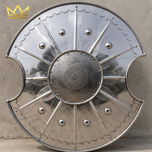 May include: A circular polished steel shield featuring a central embossed circular boss with intricate floral engravings. The shield has two symmetrical side cutouts, eight radiating raised ribs, and eight small rivets along the outer edge.